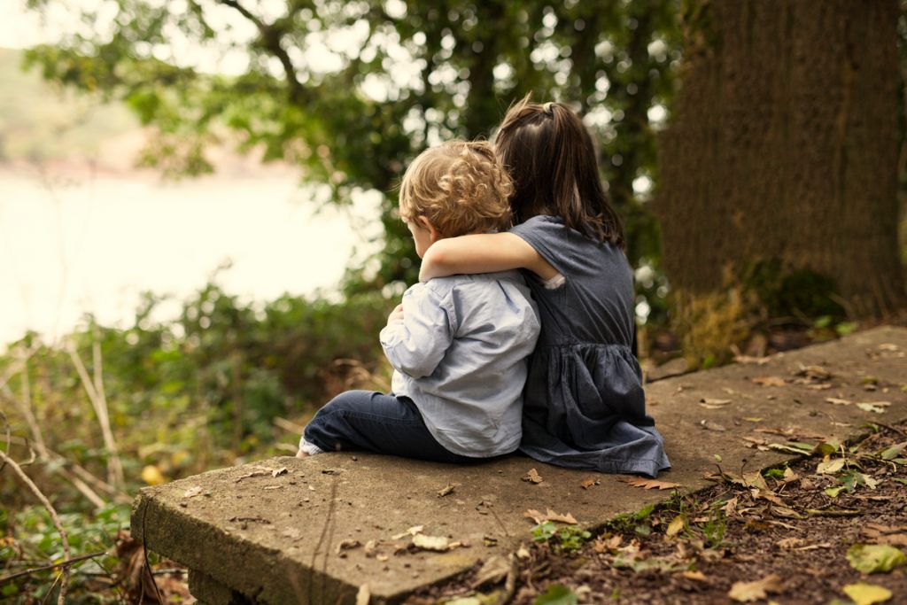Two young male and female children sitting on bench in the forest embracing and looking at the river