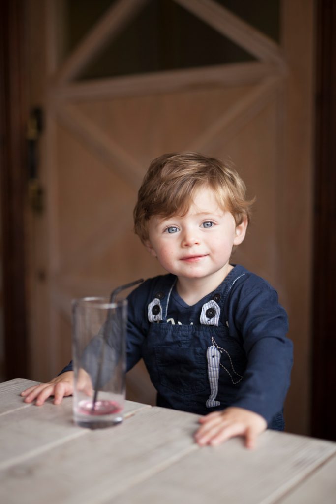 Young boy wearing a blue jumper and dungarees is supporting himself standing up against a table which has a glass on it and he is looking at the camera
