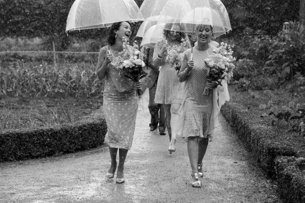 A bridal party walking on a pathway holding translucent umbrellas and bouquets of flowers while laughing